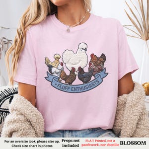 Pode incluir: Camiseta rosa claro com um desenho de v&aacute;rias galinhas e o texto "FLUFF ENTHUSIASTS" em uma faixa azul. A camiseta tem mangas curtas e gola redonda. A palavra "BLOSSOM" tamb&eacute;m &eacute; vis&iacute;vel.