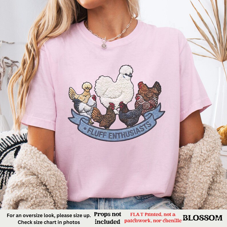 Pode incluir: Camiseta rosa claro com um desenho de v&aacute;rias galinhas e o texto "FLUFF ENTHUSIASTS" em uma faixa azul. A camiseta tem mangas curtas e gola redonda. A palavra "BLOSSOM" tamb&eacute;m &eacute; vis&iacute;vel.