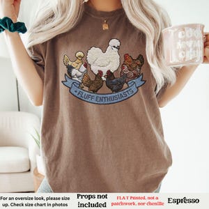 Pode incluir: Camiseta cor de caf&eacute; com uma estampa de galinhas e o texto "FLUFF ENTHUSIASTS" em uma faixa azul. A camiseta tem gola redonda e mangas curtas. A camiseta &eacute; feita de um material macio.