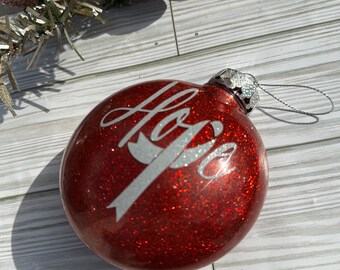 May include: A round red glitter Christmas ornament featuring the word Hope in silver script lettering with a silver ribbon graphic. The ornament has a silver metal cap and a silver hanging string attached to the top of the glass sphere.