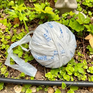 May include: A white plastic ball of plastic bags, tied together with blue tape, sits on a bed of green leaves. A small gray frog statue is visible in the background.
