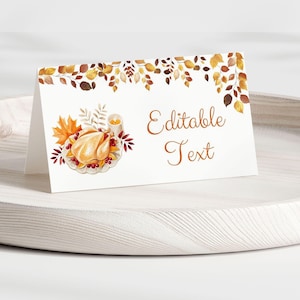 May include: A white folded place card featuring a watercolor illustration of a roasted turkey on a platter with autumn leaves and berries. The top border is decorated with fall foliage in shades of orange, yellow, and brown. The text Editable Text is printed in a brown script font.