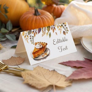 May include: A white folded place card featuring a watercolor illustration of a slice of pumpkin pie with whipped cream on a plate, decorated with autumn leaves at the top, and the black text Editable Text printed on the right side.