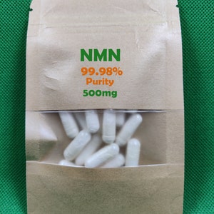Könnte beinhalten: Ein brauner Papierbeutel mit einem transparenten Fenster, in dem weiße Kapseln zu sehen sind. Auf dem Beutel steht "NMN 99,98 % Reinheit 500 mg".