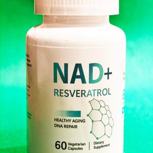 May include: White bottle with a green label that says "NAD+ RESVERATROL" and "HEALTHY AGING DNA REPAIR". The bottle contains 60 vegetarian capsules and is a dietary supplement.
