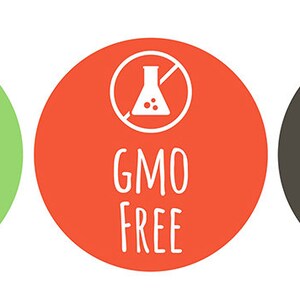 K&ouml;nnte beinhalten: Drei runde gr&uuml;ne, rote und graue Etiketten mit wei&szlig;em Text und Symbolen. Das gr&uuml;ne Etikett zeigt "VEGAN" mit einem wei&szlig;en Blattsymbol. Das rote Etikett zeigt "GMO FREE" mit einem wei&szlig;en Symbol eines Kolbens mit einem diagonalen Strich. Das graue Etikett zeigt "GLUTEN FREE" mit einem wei&szlig;en Symbol einer Weizen&auml;hre mit einem diagonalen Strich.