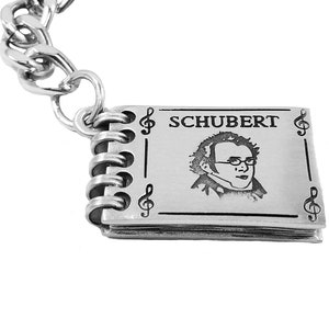 May include: Silver keychain featuring a miniature notebook with a portrait of Franz Schubert and the word "SCHUBERT" engraved on the cover. The notebook has a silver spiral binding and musical notes on the sides.