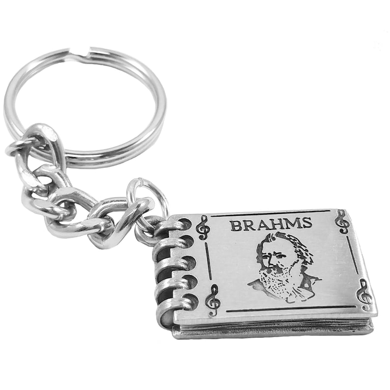 May include: Silver keychain featuring a small, engraved book with the name "BRAHMS" and a portrait of the composer. The book has a decorative, spiral binding and treble clef accents.
