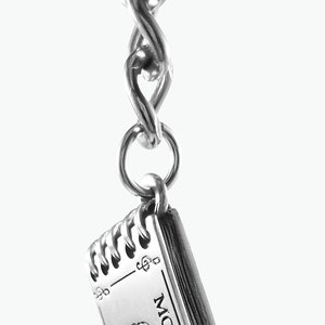 May include: Silver charm in the shape of a closed book with the word "MOZART" printed on the cover. The charm is attached to a silver chain.