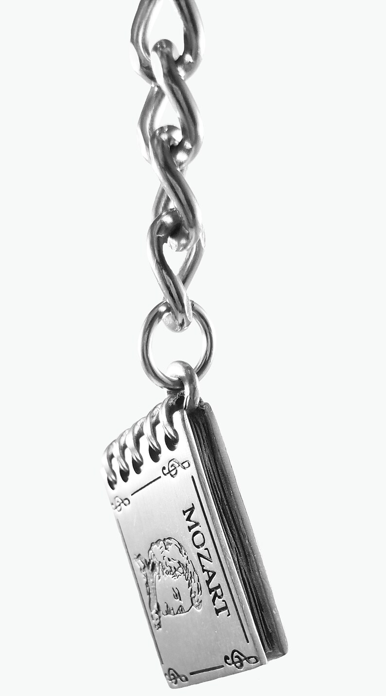 May include: Silver charm in the shape of a closed book with the word "MOZART" printed on the cover. The charm is attached to a silver chain.