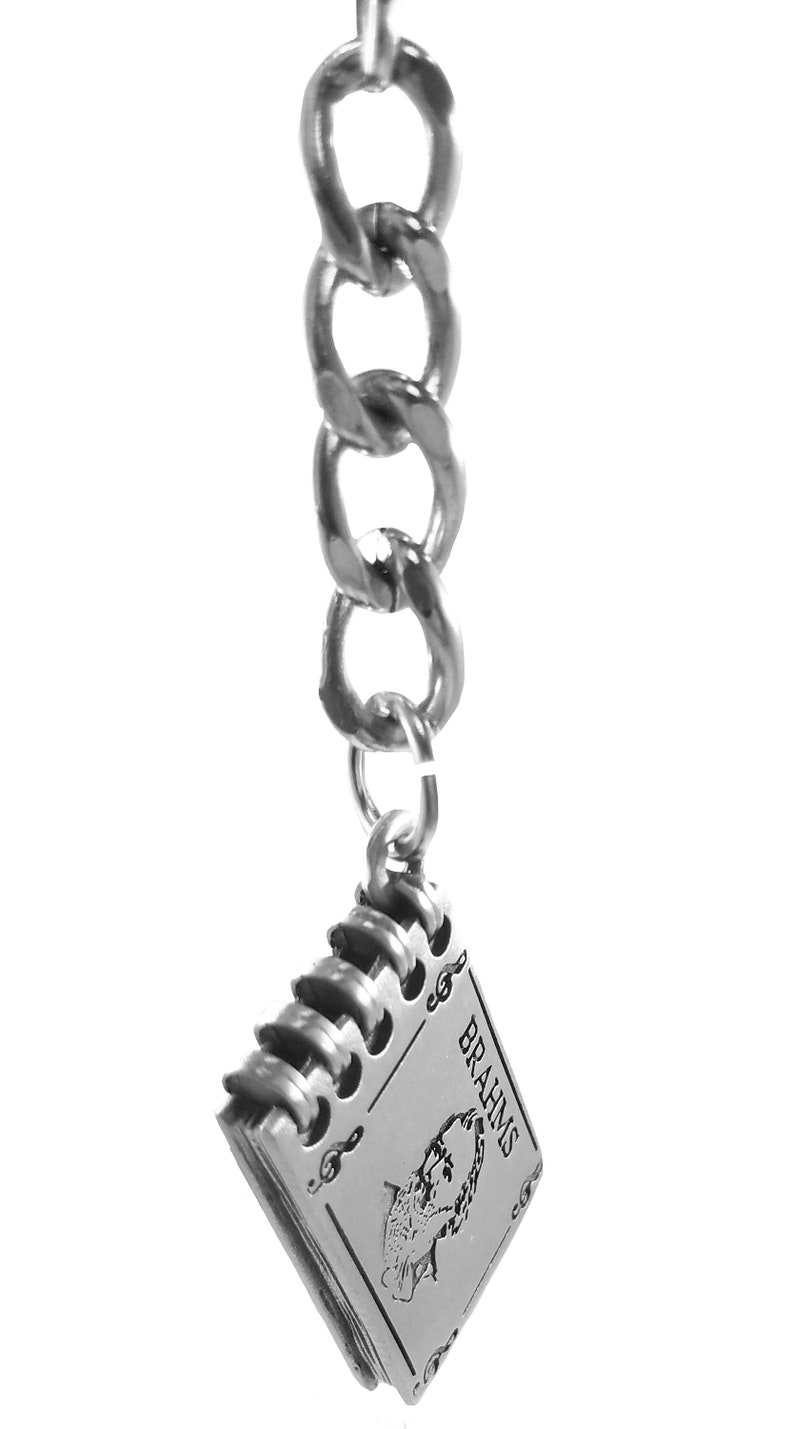 May include: A silver keychain with a silver chain and a silver charm in the shape of a book. The book has the word "BRAHMS" on the cover and a musical note design.