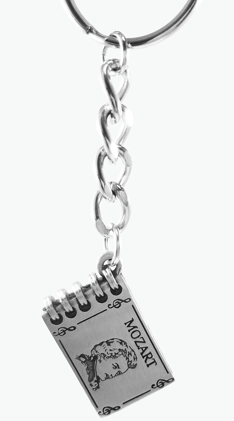 May include: Silver keychain featuring a small notebook with a portrait of Mozart and musical notes. The notebook is attached to a silver chain and a silver key ring.