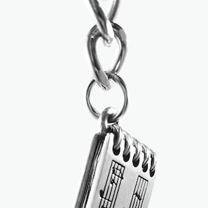 May include: Silver charm keychain featuring a small notebook with musical notes and the text "SERENADE IN G".
