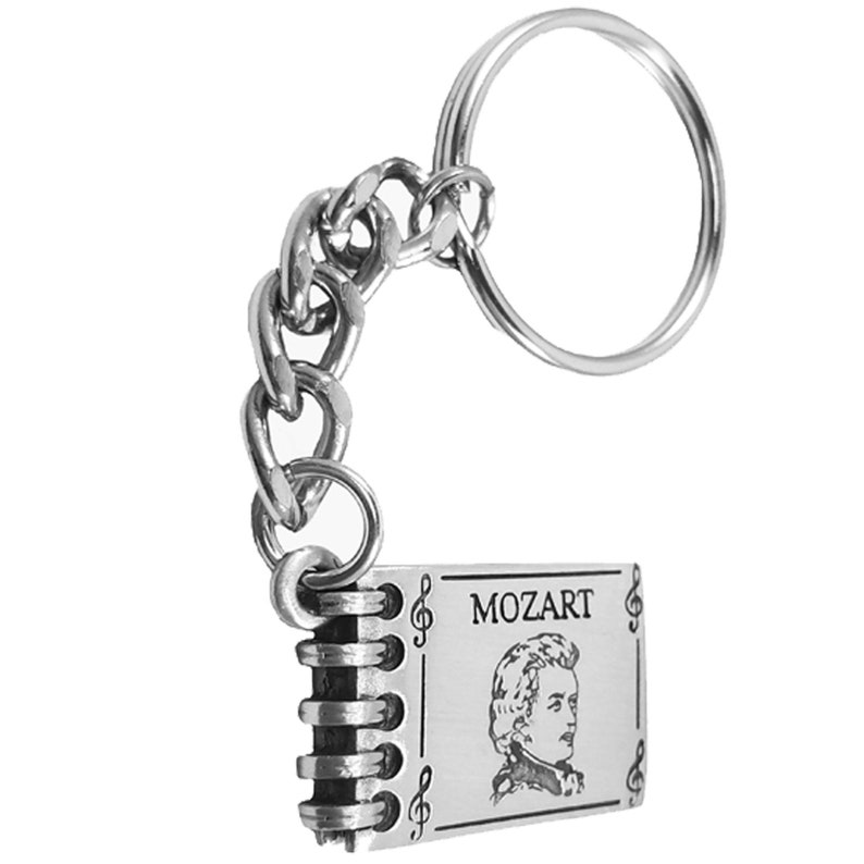 May include: Silver keychain featuring a small book with a portrait of Mozart and the word "MOZART" engraved on the front. The book has a silver chain and a silver ring.