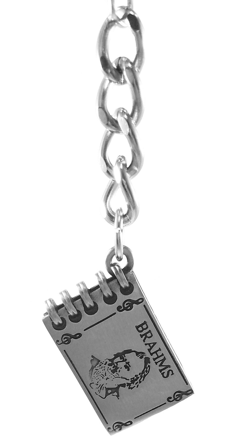 May include: A silver keychain featuring a small notebook charm with a black and white image of Johannes Brahms and the word "BRAHMS" engraved on it. The charm is attached to a silver chain.