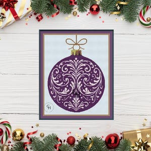 May include: A framed cross-stitch pattern of a purple Christmas bauble with white floral designs. The bauble has a gold-coloured top and a brown frame. The background is light blue. The image is surrounded by Christmas decorations.