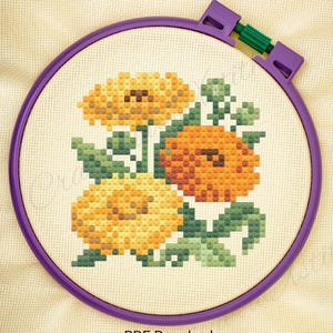 May include: A cross-stitch floral design featuring orange and yellow flowers with green leaves, set within a purple embroidery hoop. The text "PDF Download" is visible at the bottom. The design is on a cream-colored fabric.