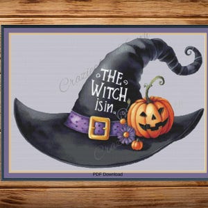 Può includere: Un disegno a punto croce incorniciato a tema Halloween. L'immagine presenta un cappello da strega nero con una fascia viola e fibbia dorata, una zucca arancione intagliata e il testo "THE WITCH is in."