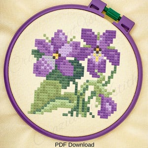 May include: A cross-stitch embroidery of purple flowers with green leaves and yellow centers, set in a purple hoop. The text "PDF Download" is printed at the bottom of the image.