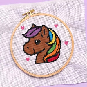 Puede incluir: Patrón de punto de cruz de un caballo marrón con una crin arcoíris y flequillo morado. El caballo está rodeado de corazones rosas sobre un fondo blanco.