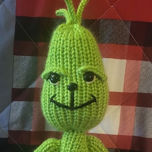 May include: A handmade, knitted Grinch doll in bright green. The doll has a distinctive face with black button eyes, a black nose, and a stitched smile. The Grinch is posed with arms at its sides, and stands on a plaid background.
