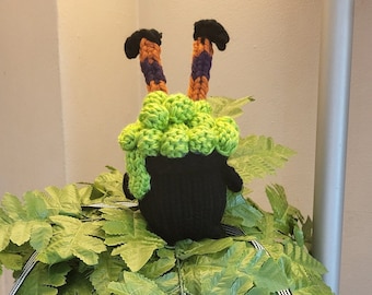 Pode incluir: Um caldeirão de bruxa preto e verde com pernas saindo do topo. O caldeirão está sentado em cima de uma planta de samambaia verde. Uma faixa preta com morcegos brancos e o texto "Trick or Treat" em letras douradas pendura abaixo da planta.
