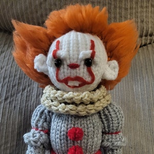 May include: A crocheted stuffed toy of Pennywise the Dancing Clown from the movie "IT". The toy is wearing a gray and red outfit with a white collar and has red pom-poms on its chest and shoes.