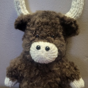 May include: A brown and white knitted plush toy shaped like a bull with white horns. The toy has black eyes and a white nose.