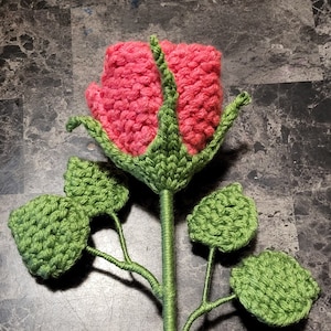 May include: A handcrafted crochet rose featuring a bright pink flower head and green stem and leaves. The petals are densely crocheted, and the stem is wrapped in green yarn. The leaves are circular and also crocheted.