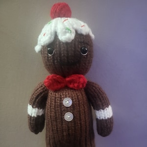 May include: A handmade, knitted gingerbread man doll. The doll is brown with white frosting on its head, a red bow tie, and white stripes on its arms and legs. It has two white buttons and a red cherry on top. The doll has black eyes.