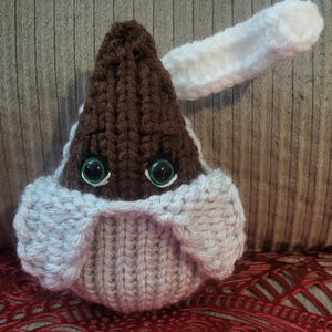 May include: A brown knitted teardrop-shaped plush toy with white knitted wings and green eyes.