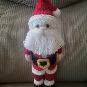May include: A crocheted Santa Claus doll with a white beard, red hat, and black boots. The doll is wearing a red and white striped suit with a black belt and a gold buckle.