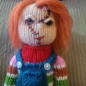 Könnte beinhalten: Handgestrickte Puppe mit orangefarbenem Haar und aufgenähten Gesichtsdetails. Die Puppe trägt einen roten Rollkragenpullover, eine blaue Latzhose mit weißen Knöpfen und einen gestreiften Pullover in Regenbogenfarben. Das Gesicht der Puppe hat aufgenähte Narben.