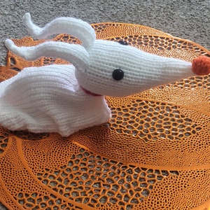 May include: A white knitted Zero plush toy from The Nightmare Before Christmas. The toy has a long snout with an orange nose, black button eyes, and a red collar. It rests on an orange, decorative, openwork placemat.