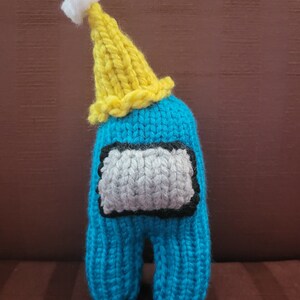 May include: A blue knitted plush toy shaped like the character from the video game Among Us, wearing a yellow knitted party hat with a white pom-pom.