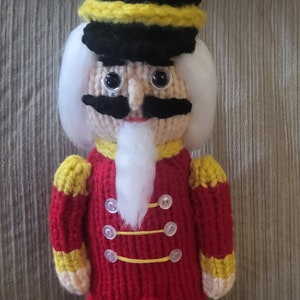 Könnte beinhalten: Eine gestrickte Nussknackerpuppe mit einer rot-gelben Uniform, schwarzen Stiefeln, einem weißen Bart und schwarzen Augen. Die Puppe trägt einen schwarzen Hut mit einem gelben Band und einer weißen Feder.