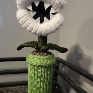 May include: A crocheted plush toy of a Piranha Plant from the Super Mario Bros. video game series. The plant is white with a black mouth and teeth, and is in a green crocheted pot.
