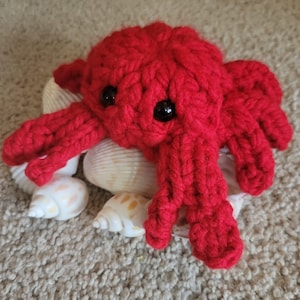 May include: A red crocheted crab toy with black eyes, sitting on a bed of white seashells.