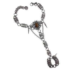 Op de afbeelding: Een zilverkleurige handkettingarmband. De armband heeft een centraal decoratief element met een bruine steen, omgeven door een bloemmotief. Spikes en kettingdetails dragen bij aan het unieke ontwerp. De armband verbindt met een ring.
