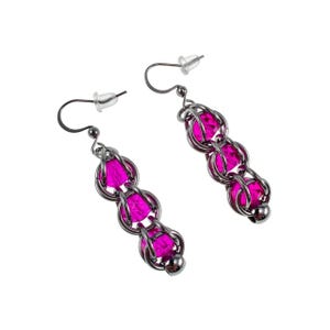 Op de afbeelding: Een paar oorhangers met drie helder roze, facetgeslepen kralen, omhuld in een zilverkleurig metalen weefsel. De oorbellen hebben een oorsteker en haaksluiting, tegen een witte achtergrond.