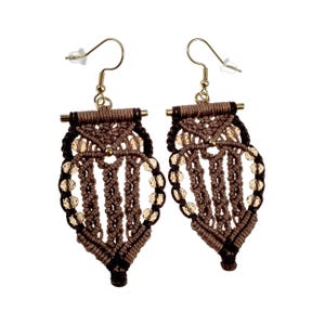 Op de afbeelding: Een paar handgemaakte macramé oorbellen. De oorbellen hebben een bruin en zwart geweven ontwerp met goudkleurige kralen. De oorbellen hebben goudkleurige oorhaken.
