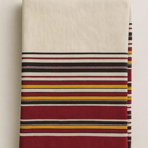 Op de afbeelding: Gevouwen crèmekleurig textiel met horizontale strepen in bordeauxrood, zwart, geel en wit. De onderste rand heeft een groene rand. Dit kan een tafelkleed, deken of decoratieve stof zijn.