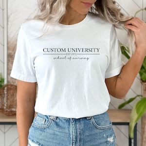 May include: A white short-sleeved t-shirt featuring black text that reads CUSTOM UNIVERSITY EST. 1972 and school of nursing in a script font, with horizontal lines separating the text, paired with blue denim jeans with a distressed hem.