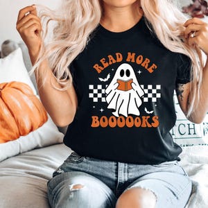 May include: A black short-sleeved t-shirt featuring a graphic of a white ghost reading an orange book. The text reads READ MORE BOOOOOKS in orange letters. The design includes a black and white checkered pattern, small stars, and bats.