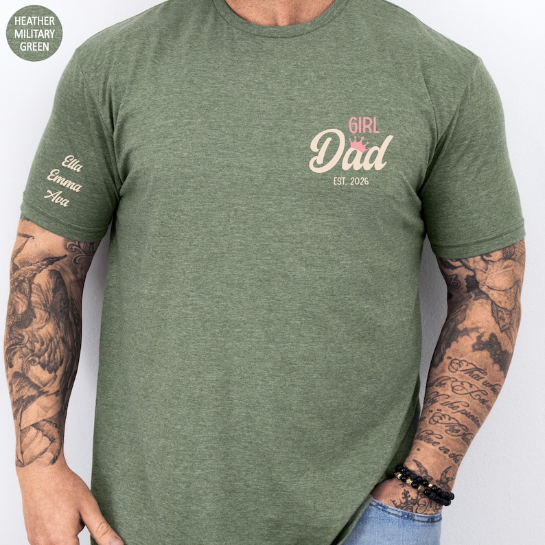 A person wearing a green t-shirt with a custom design on the left side, featuring the text "GIRL DAD EST. 2016" and the names "Ella" and "Ava".