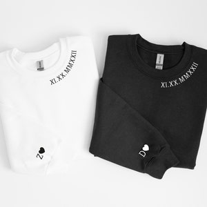 May include: Two sweatshirts, one white and one black, with a roman numeral date and a heart with a letter on the sleeve. The date is "XI.XX.MMXXII".