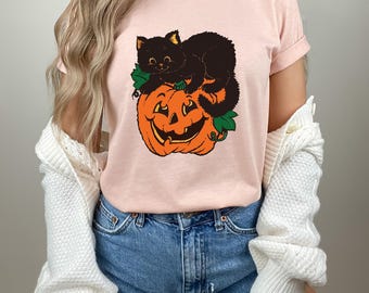 May include: A light pink short-sleeve t-shirt featuring a graphic print of a black cat sitting on top of a bright orange jack-o'-lantern pumpkin with green leaves. The shirt has a crew neckline and is worn with blue denim shorts and a white cardigan.