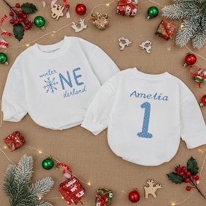 May include: Two white baby sweatshirts. One has "winter NE derland" in blue, with a snowflake. The other has "Amelia" and the number "1" in blue. Festive decorations surround the sweatshirts.