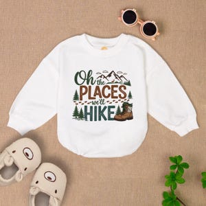 May include: White long-sleeve sweatshirt with a graphic print of mountains, trees, footprints, and a hiking boot. The text on the shirt reads "Oh the places we'll hike".
