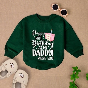 May include: Green long-sleeve sweatshirt with white text that reads "Happy First Birthday as my Daddy! Love, Ellis". A pink and white cake with a lit candle is above the text.
